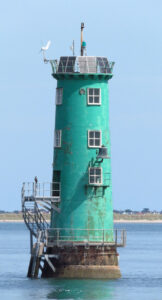 A tall turqoise-green painted beacon at sea, with several levels of windows above an external staircase and solar panels on the roof. The stone is brown near the waterline and land is a thin line in the far background behind it