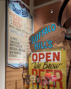 Part of the beer collection at the Smithsonian National Museum of American History in Washington DC. Behind a glass panel are taps and beautifully vintage hand-painted signage from Buffalo Bill's brewery in the 1980s. A draft beer list hangs on the wall, with prices around $2