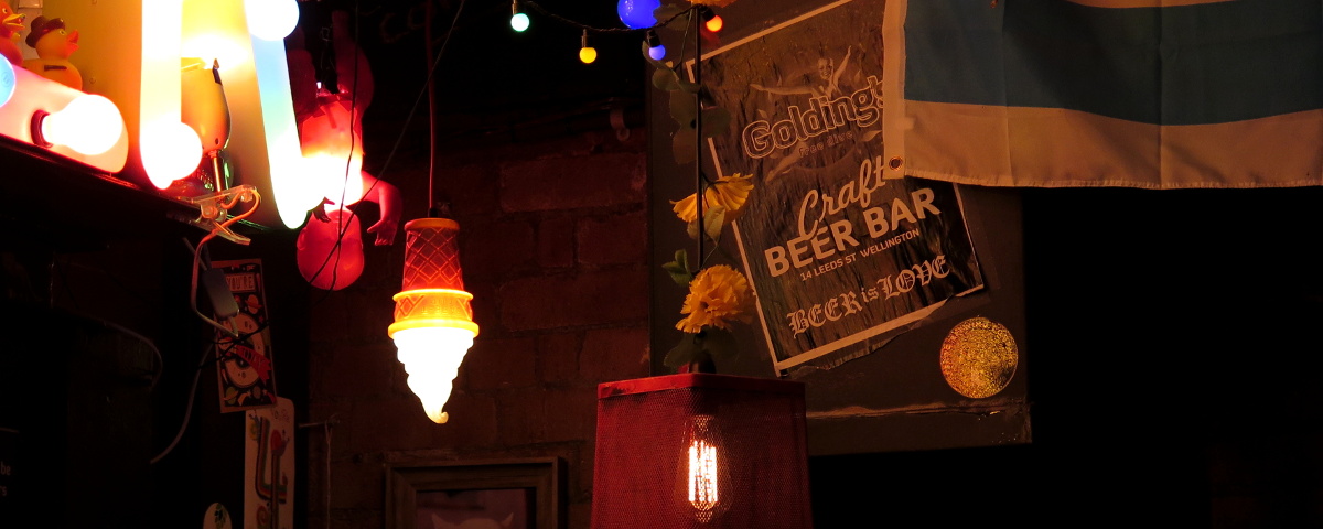 The dark corner of Golding's Free Dive in Wellington, with a poster from its opening, surrounded by colourfully light oddments in classic dive bar style; a Chicago flag, framed photo of a regular's dog, birthday cards, and a doll, a toy icecream, and a wastepaper basket all turned into hanging lamps