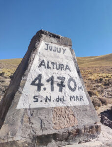 A carved altitude marker at 4,170 meters elevation in Argentina. Lettering looks hand-painted on the vaguely pyramid-shaped stone and high-desert brown vegetation indistinctly covers the still-rising slopes beyond it