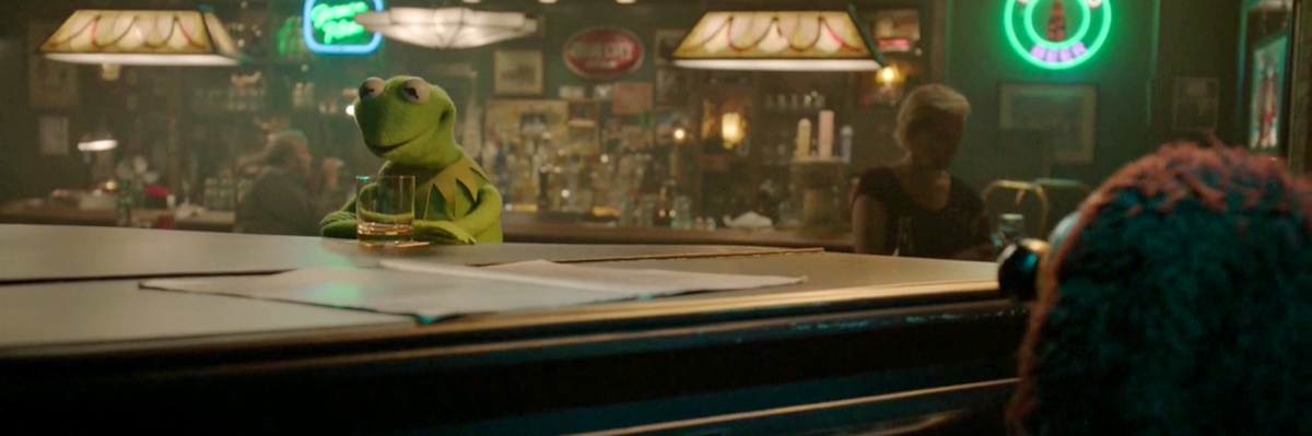 Screenshot from The Muppets (2015), episode 7. In a dark and cozy bar with neon signs on the back wall, Kermit the frog sits, arms crossed, with a glass of whiskey at the piano where Rowlf the dog is playing