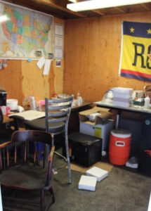 A somewhat blurry photo as if from an old and/or dirty phone camera, looking through the door into a messy office. Mismatched wooden chairs are in front of disorganised desks, with boxes on the floor. A map of the U.S. with various pieces of paper pinned to it, and a large flag with the Rogue logo are on the otherwise bare wooden walls.