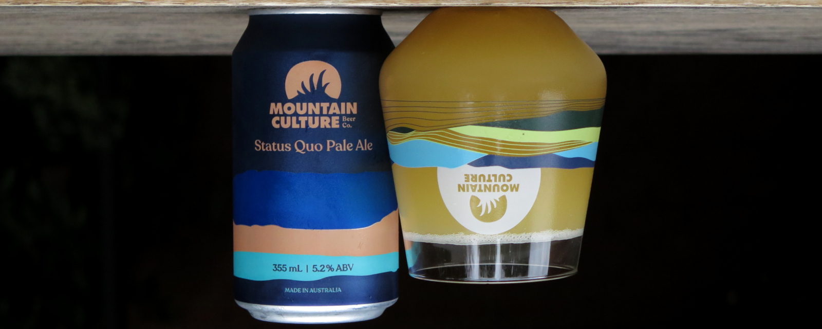 A can of Mountain Culture Status Quo hazy pale next to a branded glass full of the same. The can was upside down but then the entire photo is rotated so the glass appears upside down with the beer mysteriously levitating inside it while both seem to float in a dark void.