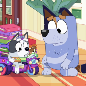 Screenshot from the cartoon Bluey, episode 'Library' (s02e30), depicting the interior of a house where Stripe (a blue heeler dog), driving, is kneeling down to talk to his daughter Muffin (a grey and white heeler dog in a tiara), who is riding a toy bike among a pile of books