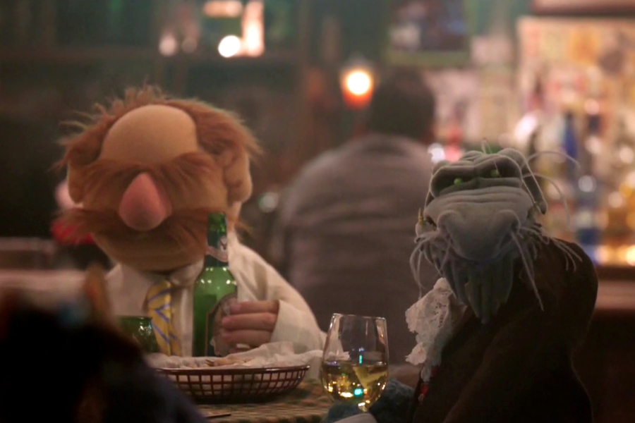 Screenshot from The Muppets (2015) episode 4. Swedish Chef and Uncle Deadly are sitting at a table in Rowlf's Tavern, Chef has a green bottle lager (in his human hand) and Deadly is staring into the camera while holding a glass of white wine