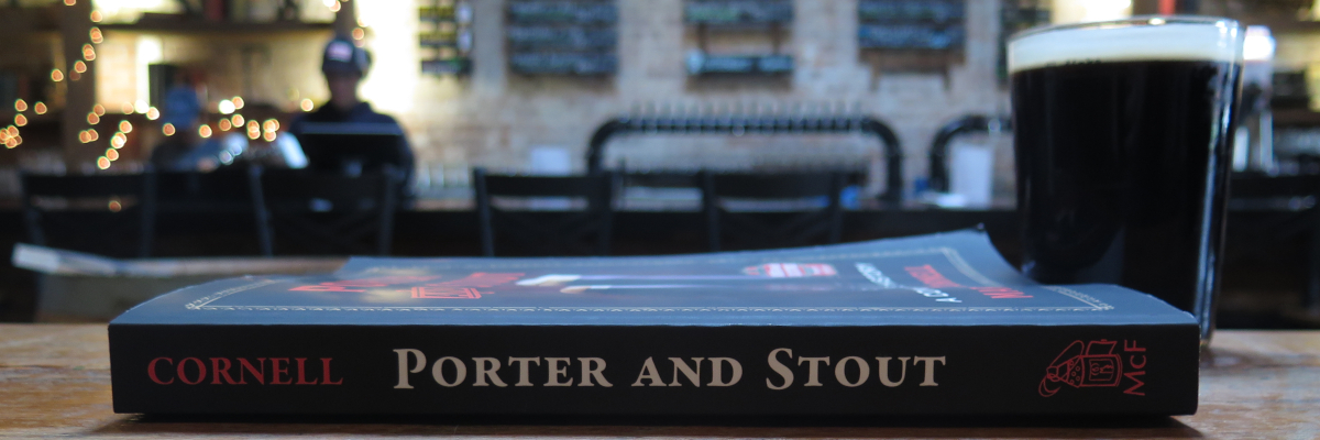 Martyn Cornell's 'Porter and Stout' book laying on its side on a wooden table with a glass of dark beer beside it (spoiler alert: neither a porter nor a stout) in a brewery taproom with the bar blurrily visible in the background