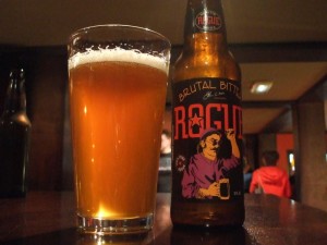A glass of orange-gold beer in a straight-sided 'Boston' pint, next to its bottle. The black label reads Rogue Brutal Bitter and there's a cartoon image of a man with a beer in one hand and the other raised in a fist.